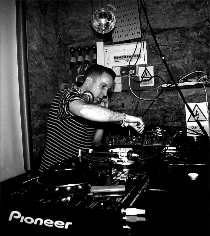 Fotografía en blanco y negro de Andrew Weatherall inclinado sobre unos platos y una mesa de mezclas, con los auriculares colgando junto a la oreja, en una sala de paredes de piedra con una bola de espejos en el techo.