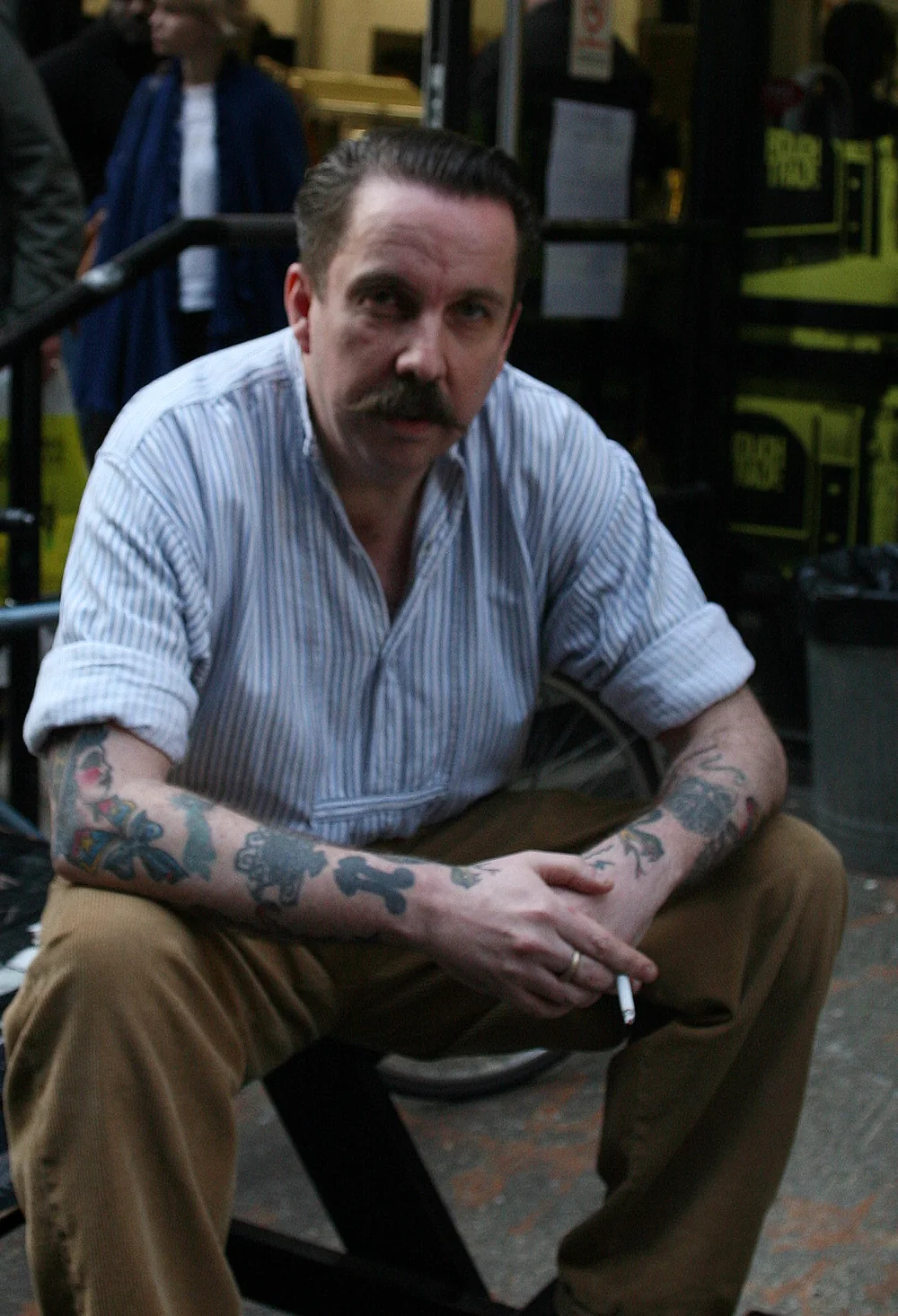 Andrew Weatherall sentado en la calle, con bigote y camisa de rayas azules con las mangas remangadas, los antebrazos tatuados y un cigarrillo entre los dedos, mirando a cámara.