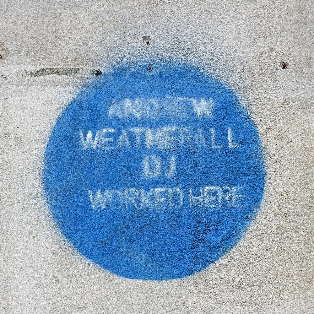 Círculo azul pintado con aerosol sobre una pared clara, con la inscripción «ANDREW WEATHERALL DJ WORKED HERE» en letras blancas, parcialmente desgastada.