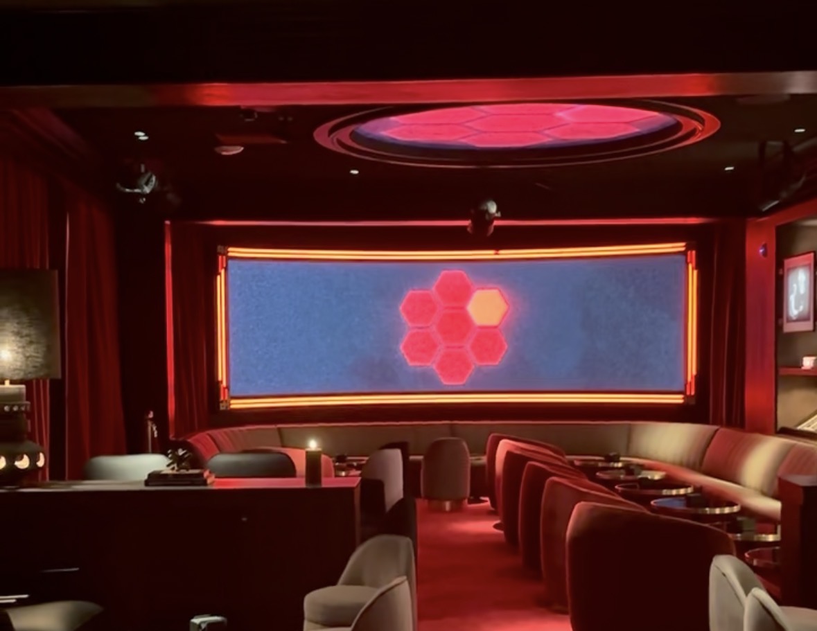 Sala a oscuras con luz roja: al fondo, una gran pantalla rectangular con siete hexágonos; encima, un óculo en el techo; delante, sofás y butacas.