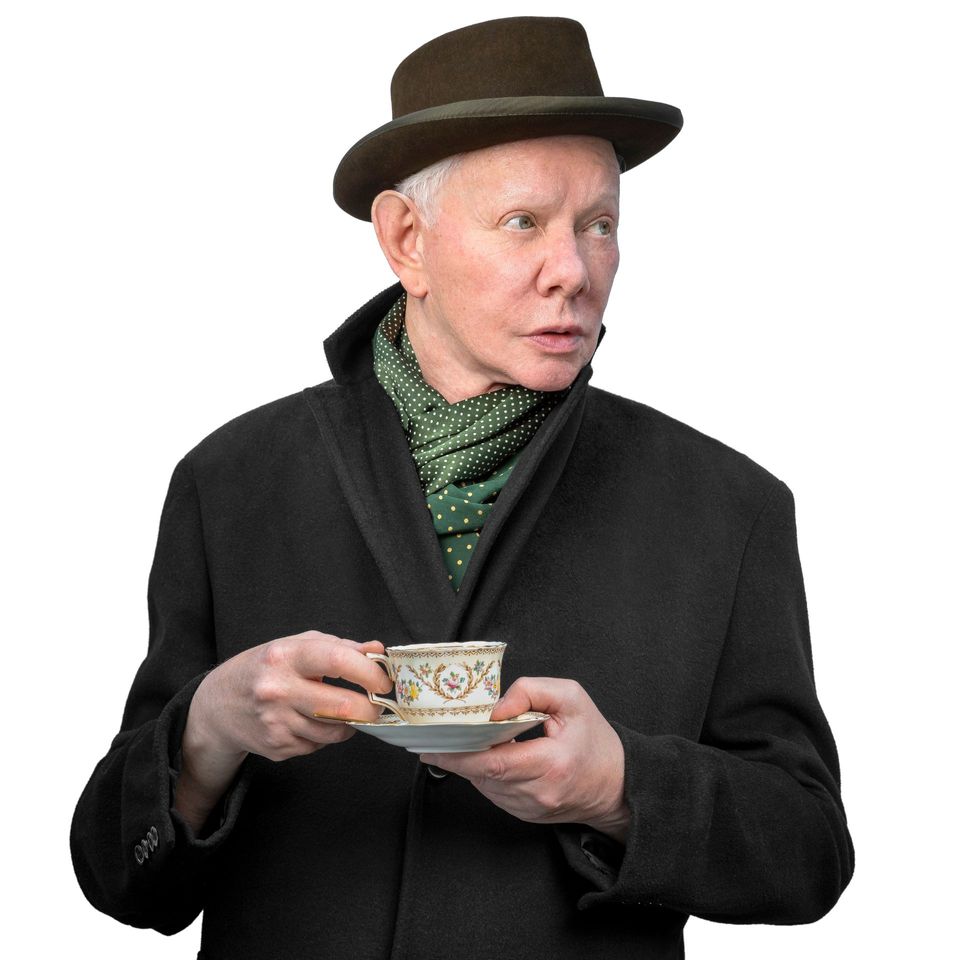Joe Jackson con sombrero verde oscuro, bufanda verde de lunares y abrigo negro, con una taza de porcelana y su platillo en las manos, sobre fondo blanco.
