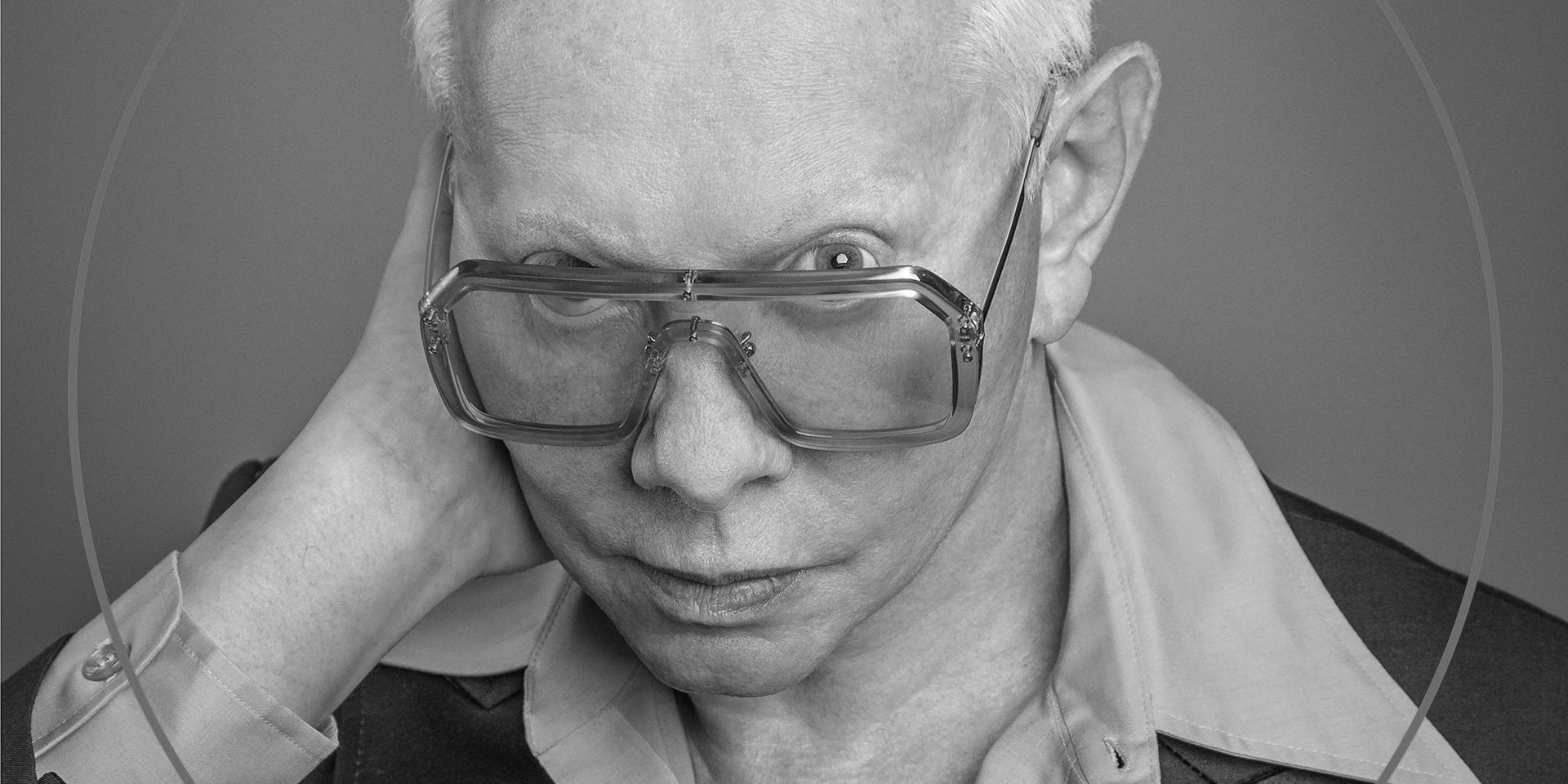 Primer plano de Joe Jackson con unas grandes gafas de lentes tintadas y la mano detrás de la oreja, sobre fondo gris con un círculo fino.