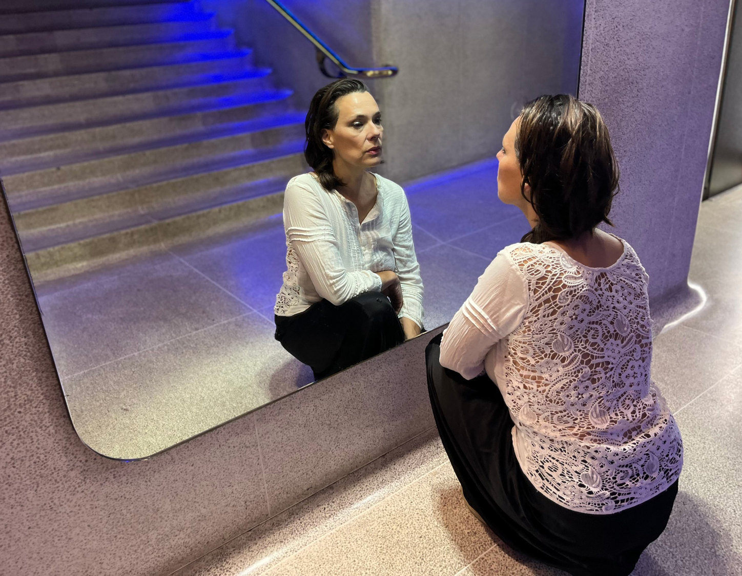 Laetitia Sadier en cuclillas ante un espejo, en una escalera iluminada de azul: se ve su reflejo y, en primer plano, su espalda con una blusa blanca de encaje.