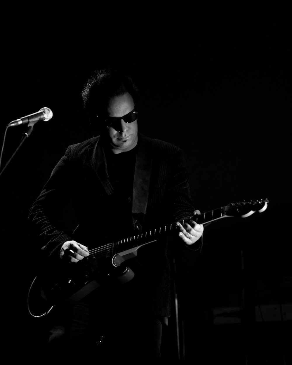Sean “Grasshopper” Mackowiak, wearing sunglasses, plays an electric guitar next to a microphone, in black and white, against an almost black background.