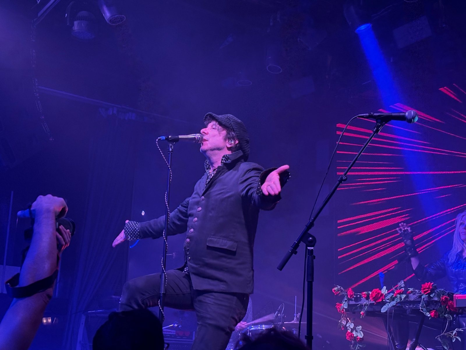 A concert in blue and violet light: a man in a cap sings into a microphone with his arms open; in the foreground, on the left, the hand of someone holding a camera.