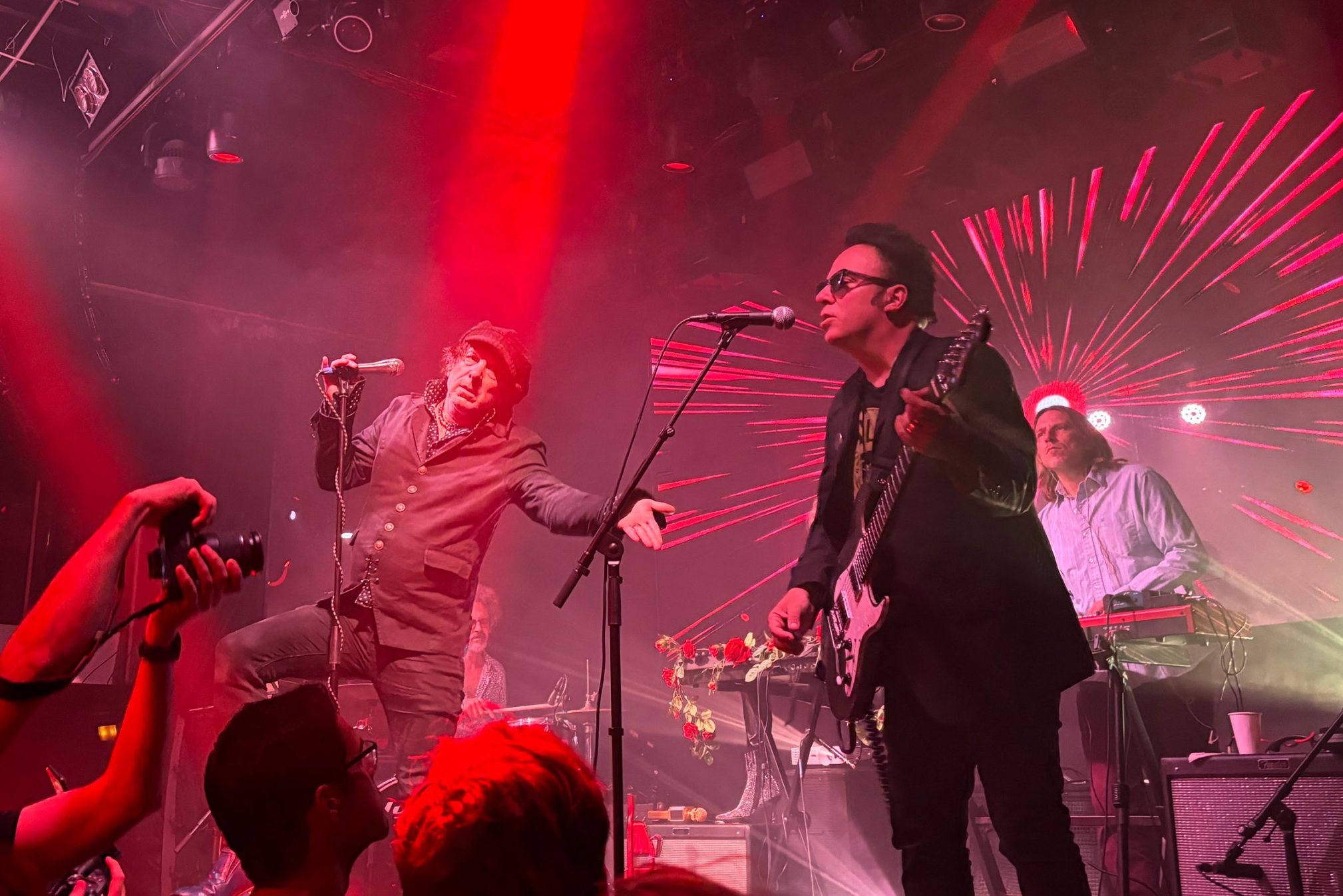 A concert in red light: two musicians at their microphones, a keyboard player at the back and, in the foreground, audience members holding a camera.