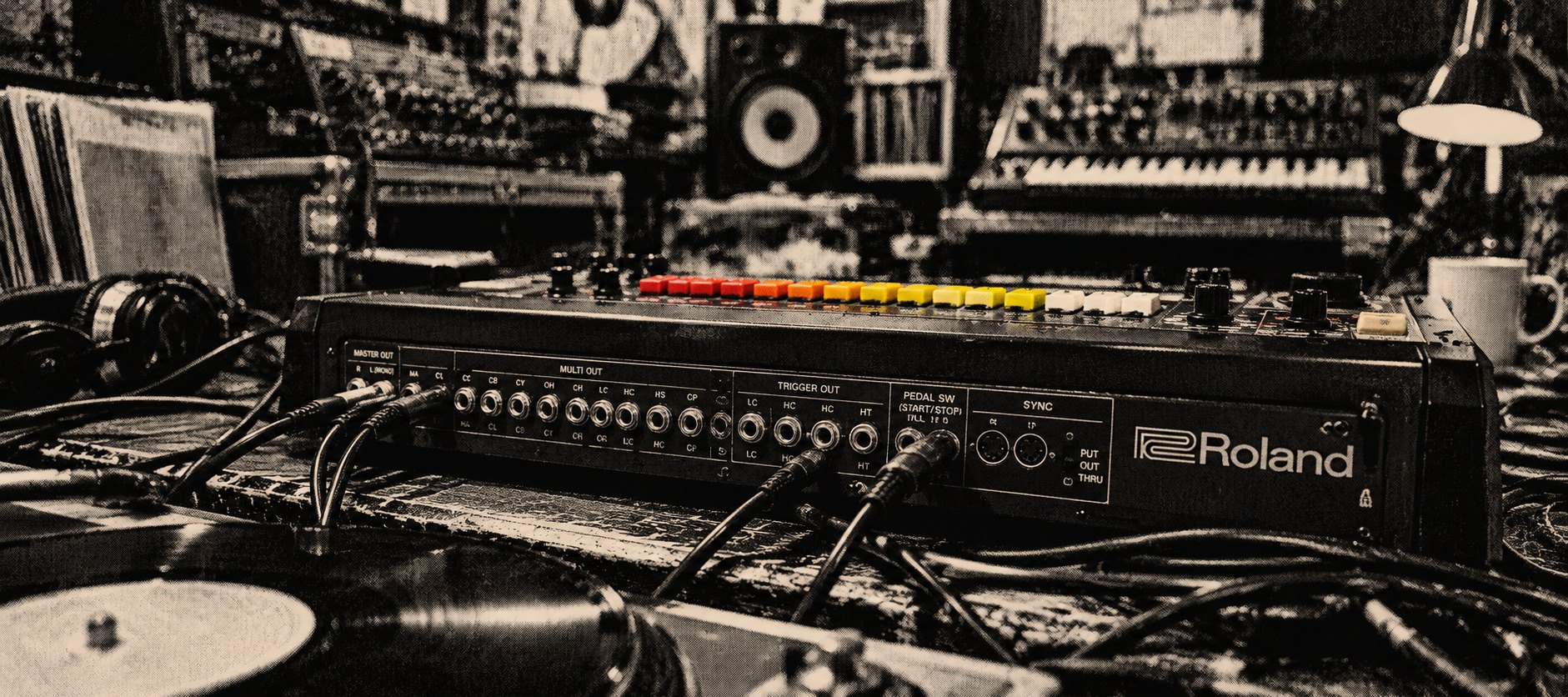 Panel trasero de una caja de ritmos Roland con sus entradas y salidas, varios cables conectados y, detrás, sus botones de colores; alrededor, un tocadiscos, vinilos y un estudio lleno de equipos.