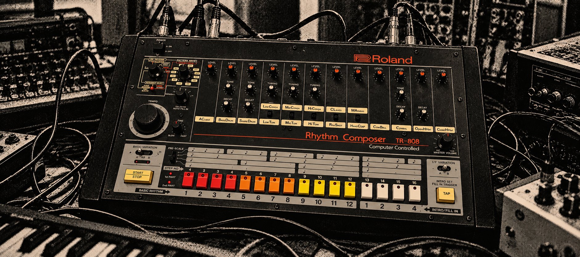 Una Roland TR-808 vista de frente, con sus botones de colores en rojo, naranja, amarillo y blanco, rodeada de cables, teclados y sintetizadores, en blanco y negro con tono sepia.
