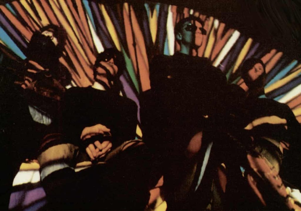 Cuatro integrantes de Spacemen 3, vistos desde abajo, delante de una proyección psicodélica de rayas de colores.