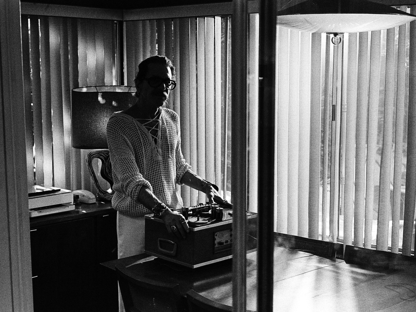William Basinski, en blanco y negro, de pie junto a una grabadora de cinta abierta sobre una mesa, ante persianas verticales y una lámpara colgante.